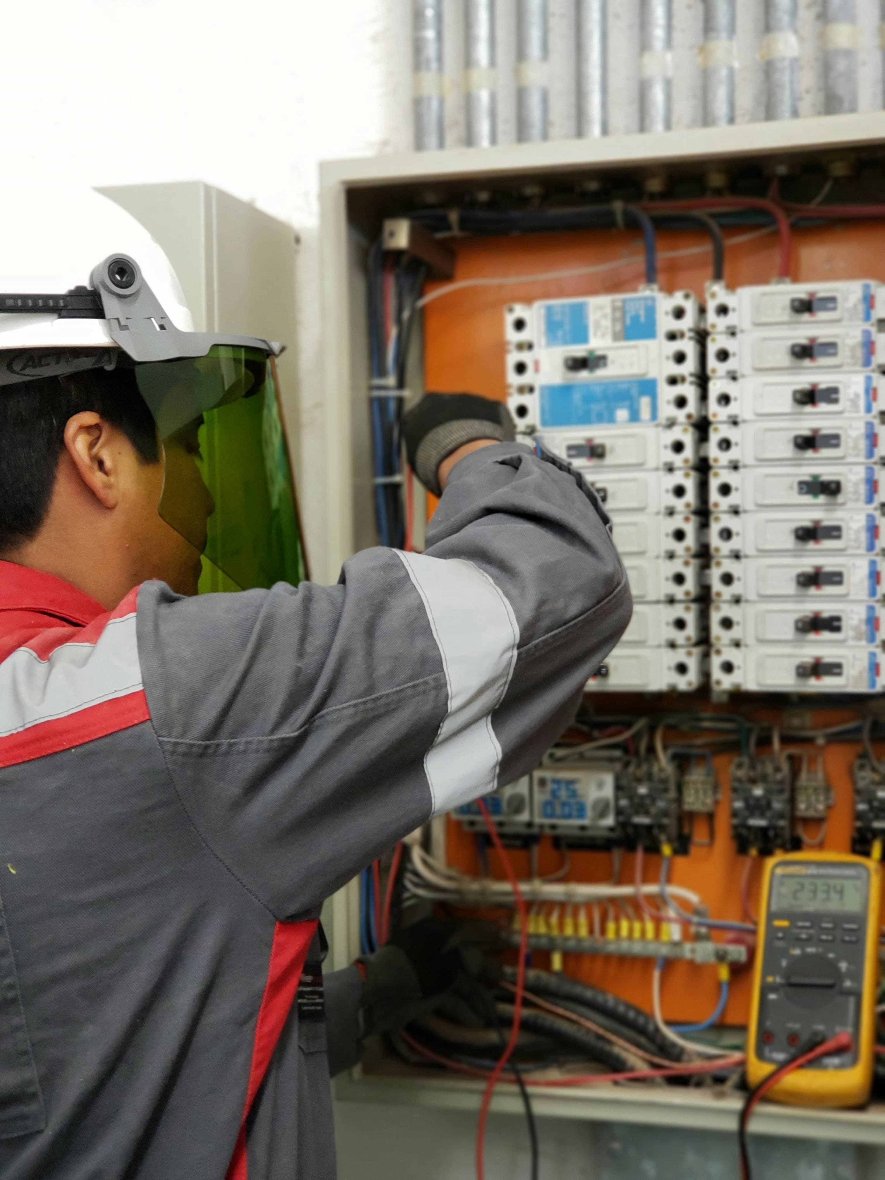 Electrician in a hard hat and face shield testing an electrical panel